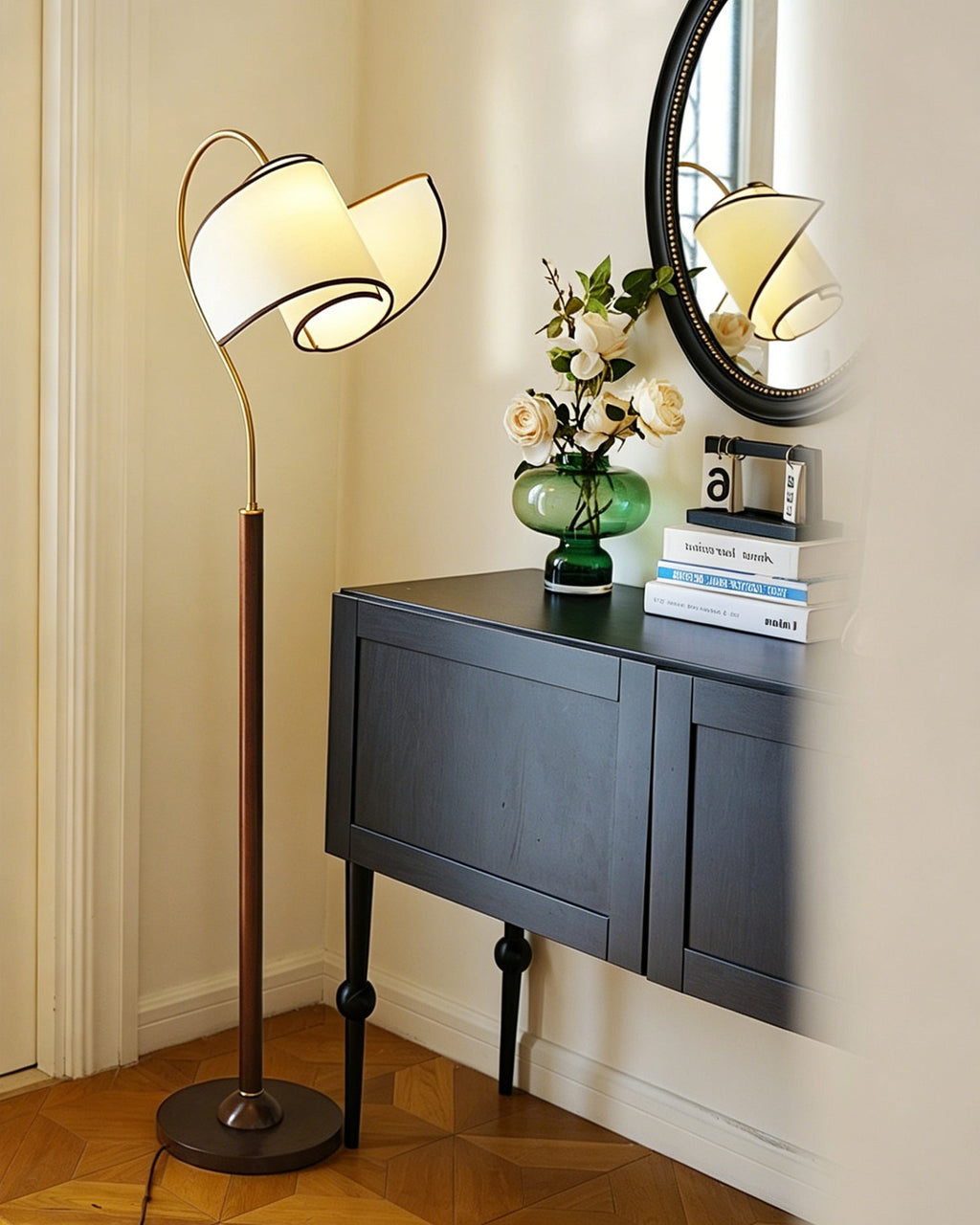 A Clérachic floor lamp with a looped white shade stands beside a black cabinet, a green glass vase and a round mirror.