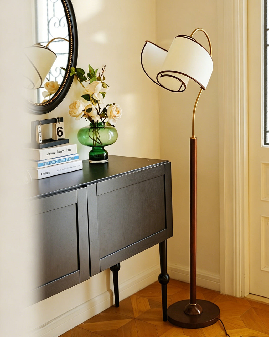 A Clérachic arched floor lamp beside a dark console with white flowers in a green vase, in a bright, panelled hallway.