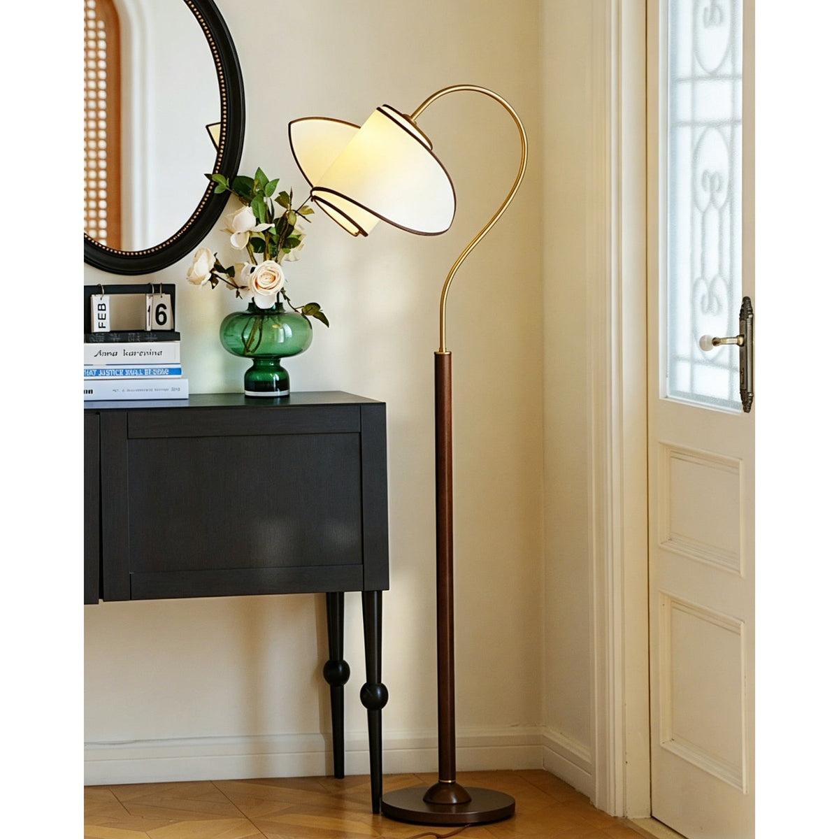 A Clérachic floor lamp stands near a black cabinet and a white door with a glazed panel, its shade arching over the cabinet.
