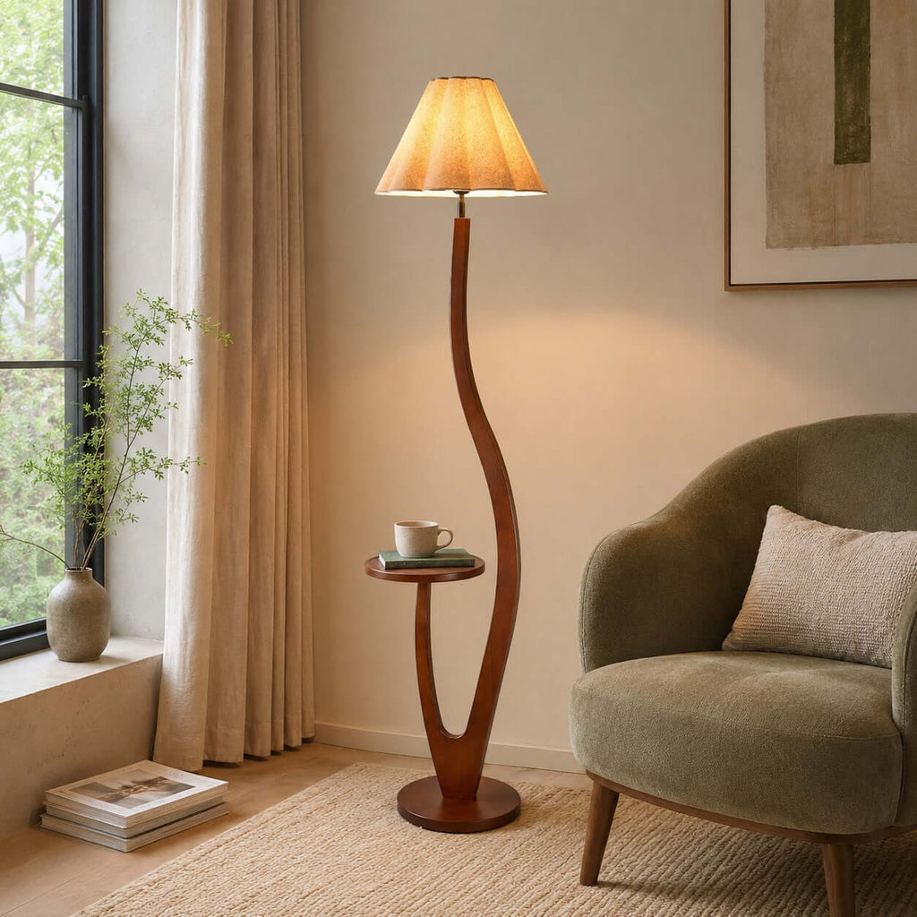 Warm light glows through a pleated shade above a curved wooden stem with a small tray holding a cup, beside a green armchair, from Clérachic.