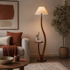A curved walnut-toned stem and round side tray stand beside a grey sofa with a rust throw, topped with a cream tapered shade, on this Clérachic floor lamp.