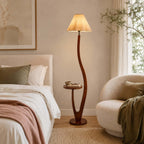Beside a bed with white and rose bedding, a Clérachic floor lamp stands on its round wooden base with a pleated shade lit warm.