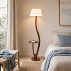 A rounder, textured white shade sits on this Clérachic floor lamp beside a bed with blue bedding, a glass of water on its tray.