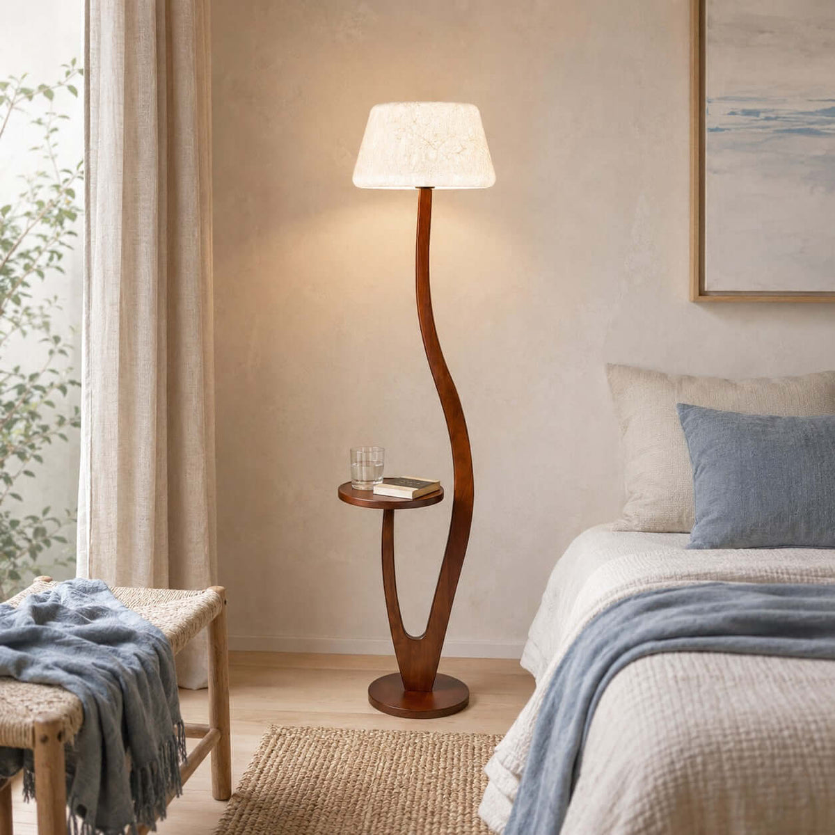 A rounder, textured white shade sits on this Clérachic floor lamp beside a bed with blue bedding, a glass of water on its tray.