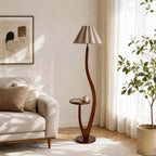 A pleated taupe shade, unlit, sits above this Clérachic floor lamp's curved stem and tray, beside a cream sofa and a leafy plant.