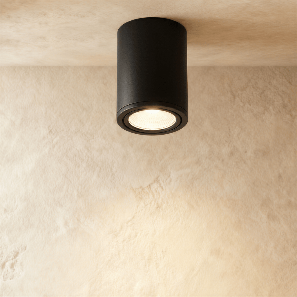 Clérachic black aluminium cylinder downlight, surface-mounted on a limewash ceiling and glowing warm from its recessed lens.