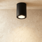 Clérachic black aluminium cylinder downlight, surface-mounted on a limewash ceiling and glowing warm from its recessed lens.
