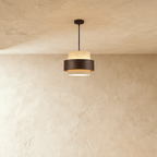 Clérachic small drum pendant of speckled travertine banded in dark wood, hung from a slim black rod against a limewash wall.