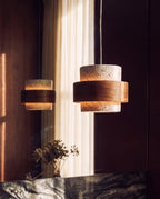 Clérachic travertine drum pendant with a wood band glowing warm, its reflection showing in a mirror above a dark marble ledge.