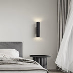 Clérachic black textured cylinder wall light above a bedside table, casting soft light up and down a pale wall.