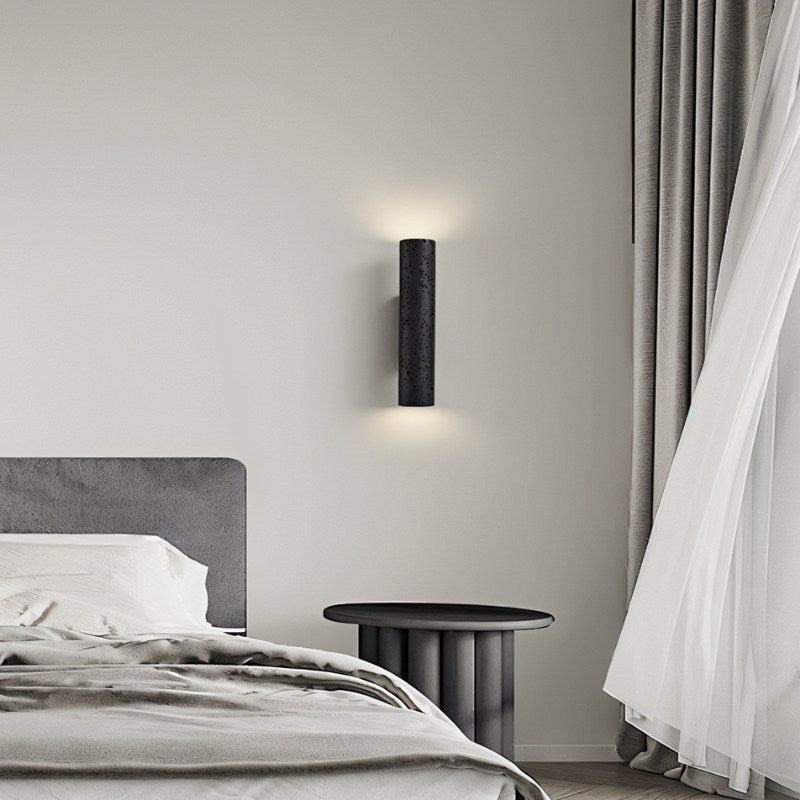 Clérachic black textured cylinder wall light above a bedside table, casting soft light up and down a pale wall.