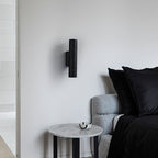Clérachic black perforated-texture cylinder wall light on a white wall beside a grey sofa and a round marble side table.