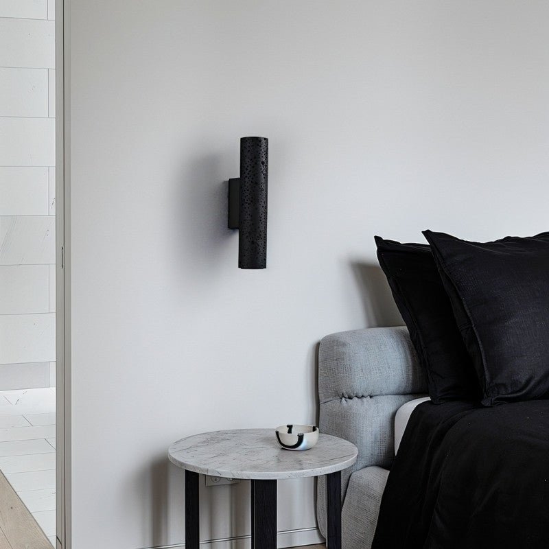 Clérachic black perforated-texture cylinder wall light on a white wall beside a grey sofa and a round marble side table.