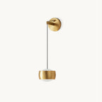 Brass Clérachic wall light on a plain white backdrop, a rounded cylinder shade hanging from a thin black cable and a round backplate.