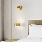 Brass Clérachic wall light glowing beside a bed with white pillows, a wood-slat headboard and a curtain at the left.