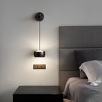Black Clérachic wall light lit above a black bedside table, with a grey padded headboard and a triple wall switch beside it.
