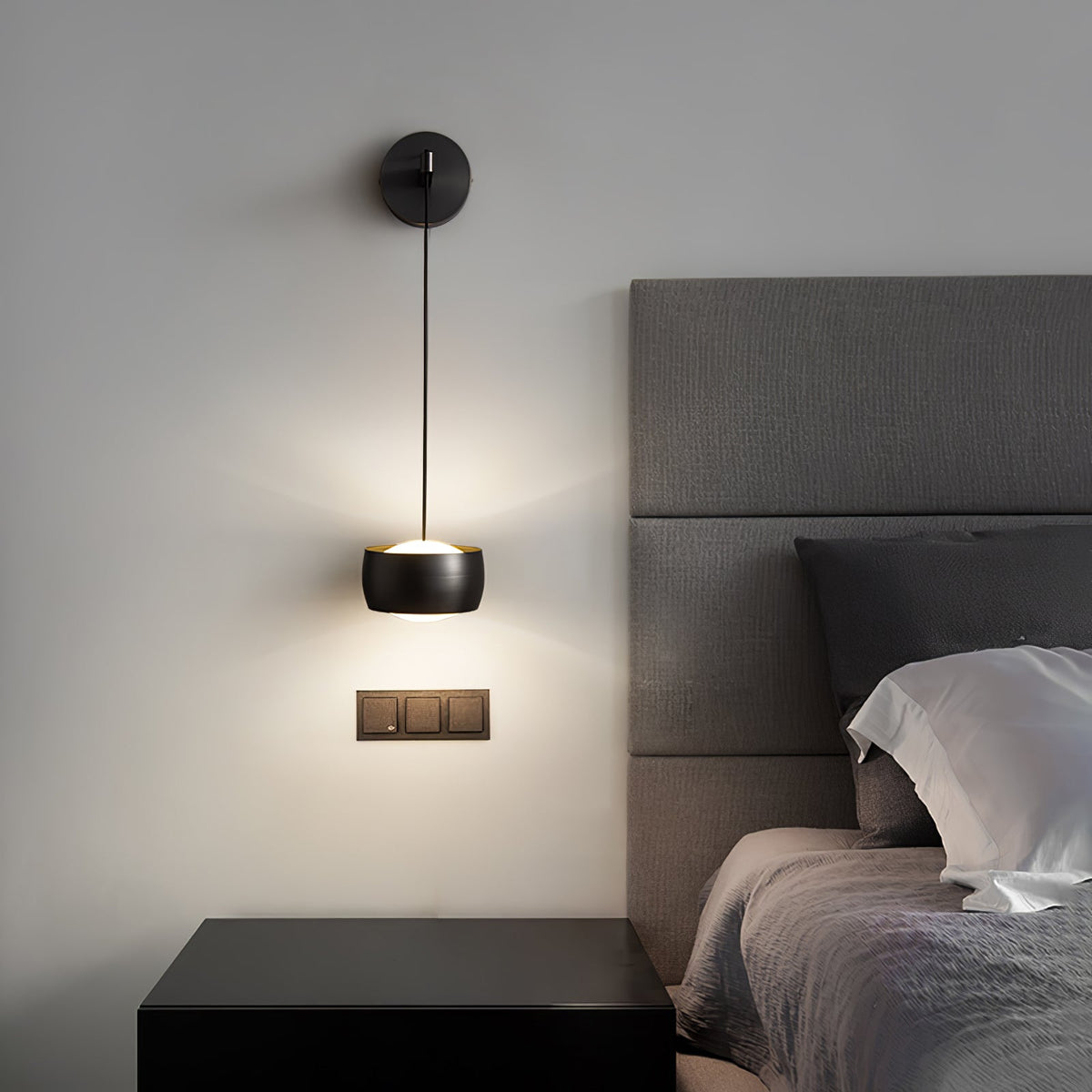 Black Clérachic wall light lit above a black bedside table, with a grey padded headboard and a triple wall switch beside it.