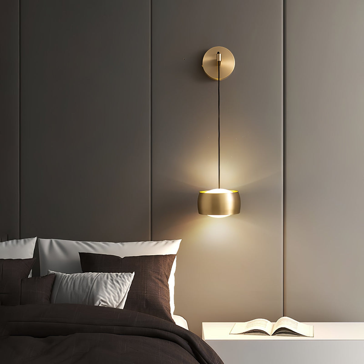 Lit brass Clérachic wall light on a taupe panelled wall above a bed with brown cushions and an open book on the shelf.
