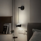 Lit black Clérachic wall light by a bed, with a shelf of books, a dried flower stem and wall sockets below.