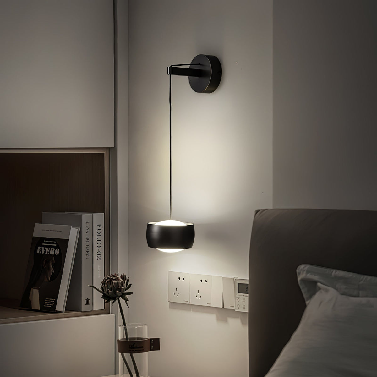 Lit black Clérachic wall light by a bed, with a shelf of books, a dried flower stem and wall sockets below.