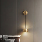 Brass Clérachic wall light throwing light up and down a taupe wall, with brown and grey pillows at the foot of the frame.