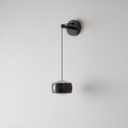 Black Clérachic wall light on a light grey wall, its glossy cylinder shade hanging below a round backplate on a thin cable.