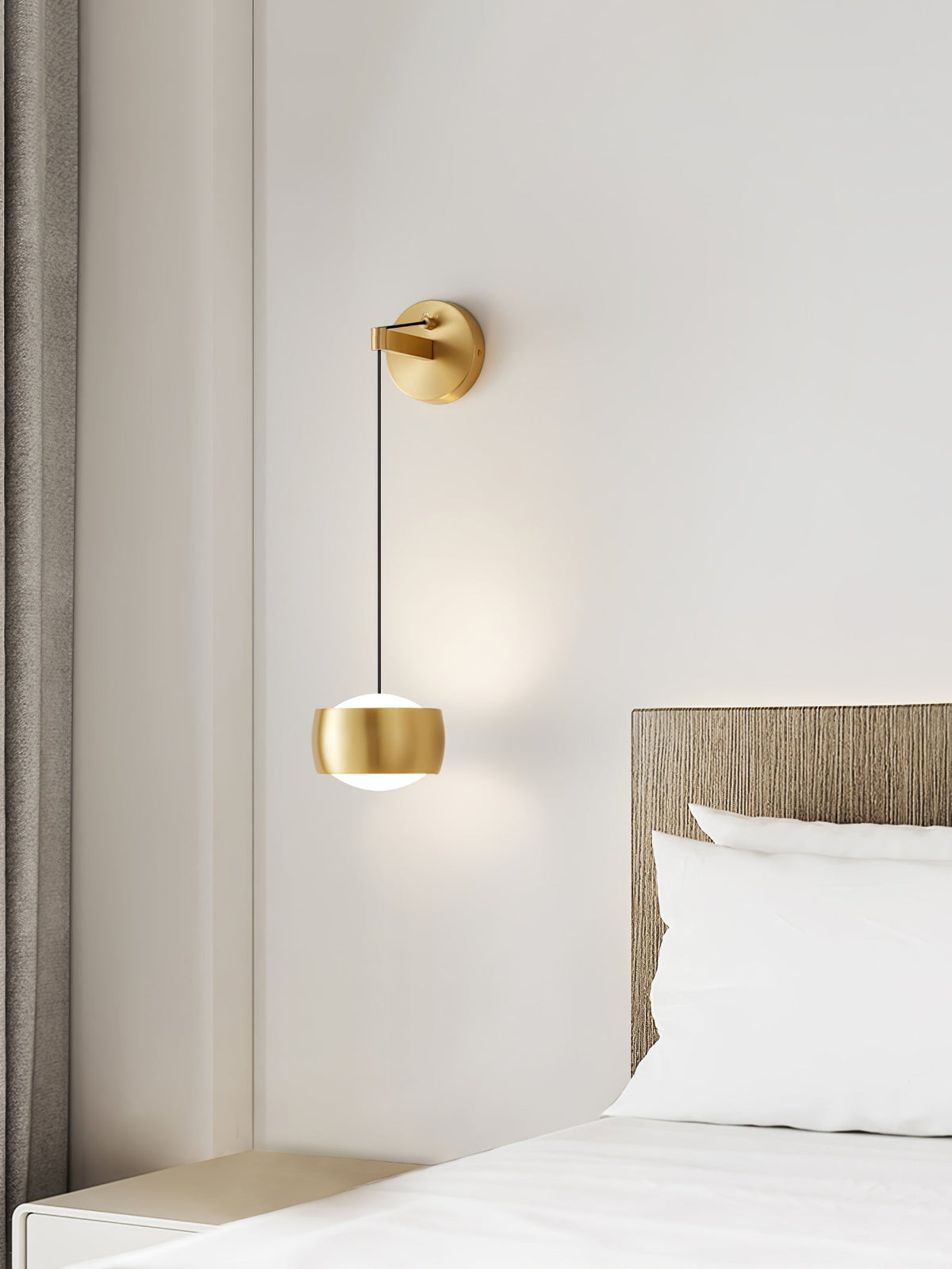 Small brass Clérachic wall light lit beside a window and a bed with white pillows, against a pale wall.