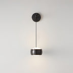 Front view of a black Clérachic wall light on a light grey wall, its shade lit white underneath and a round backplate above.