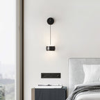 Black Clérachic wall light on a white wall by a floor-to-ceiling window, above a dark bedside cabinet and wall sockets.