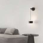 Black Clérachic wall light on a pale wall above a grey sofa and a smoked-glass side table.