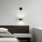 Lit black Clérachic wall light above a bed with a dark wood headboard and a pale floating bedside shelf.