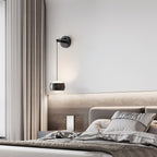 Black Clérachic wall light over a bed with grey linen and a lit wood-grain headboard panel, with a curtain at the left.