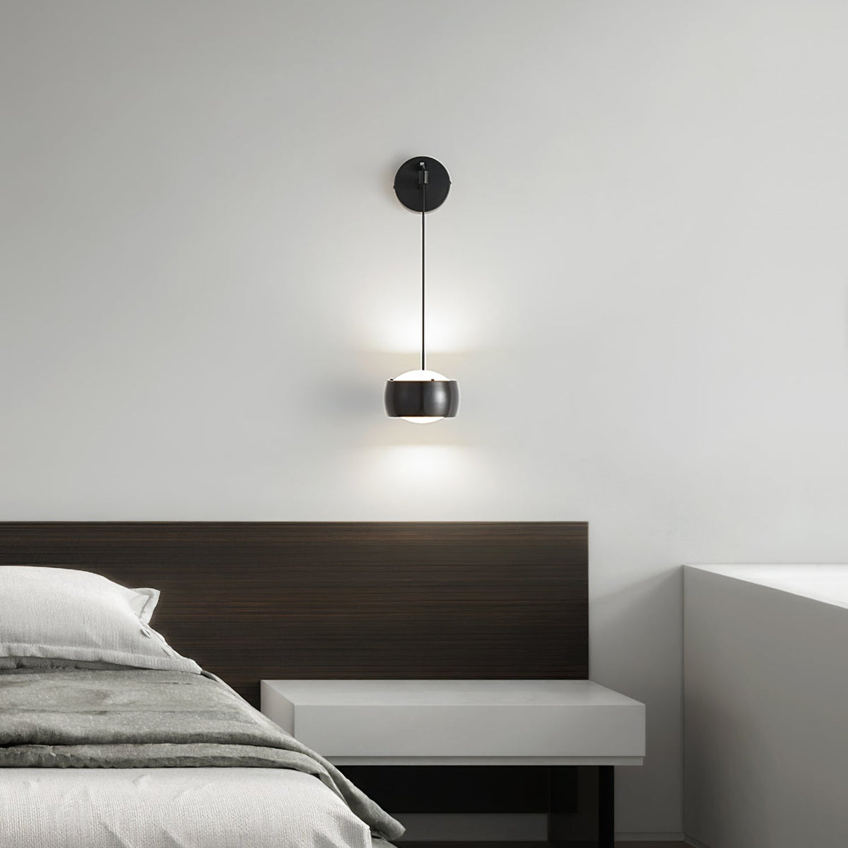 Close view of a lit black Clérachic wall light above a bed with a dark wood headboard and a pale shelf.