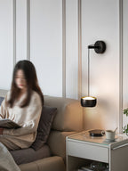 Black Clérachic wall light lit beside a sofa where a person reads, with a white bedside table and a cup and saucer.