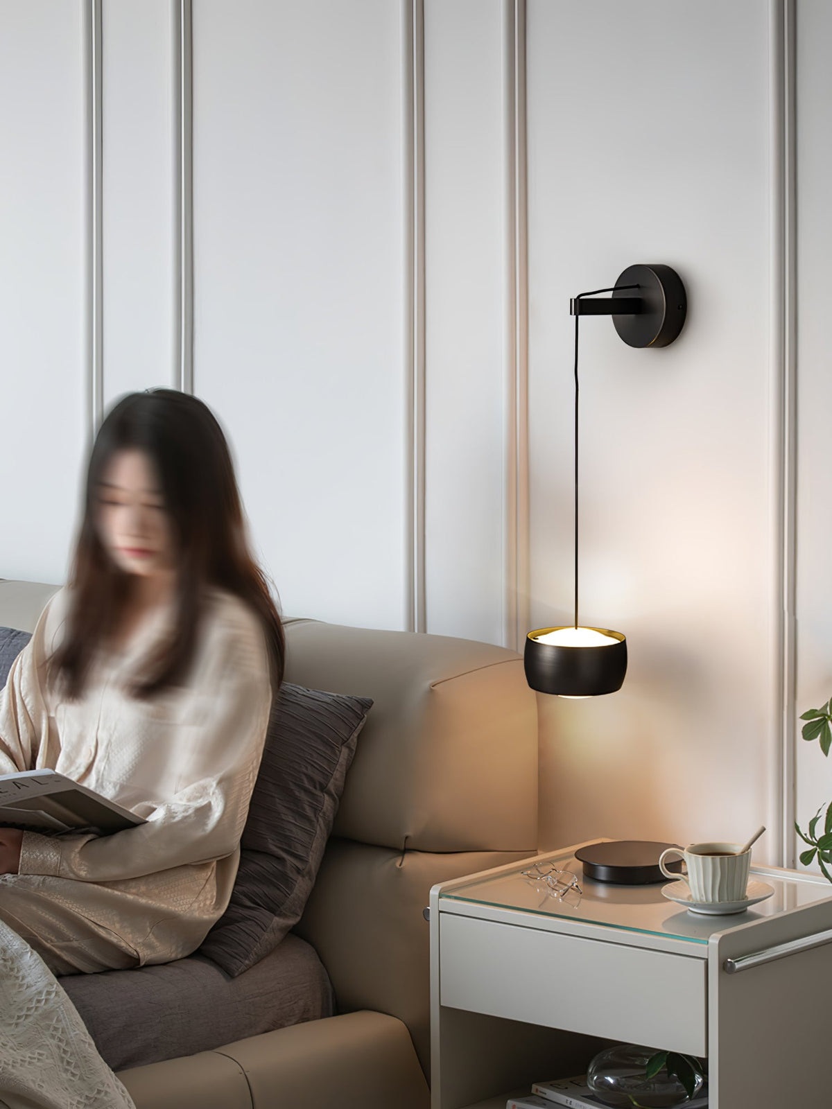 Black Clérachic wall light lit beside a sofa where a person reads, with a white bedside table and a cup and saucer.