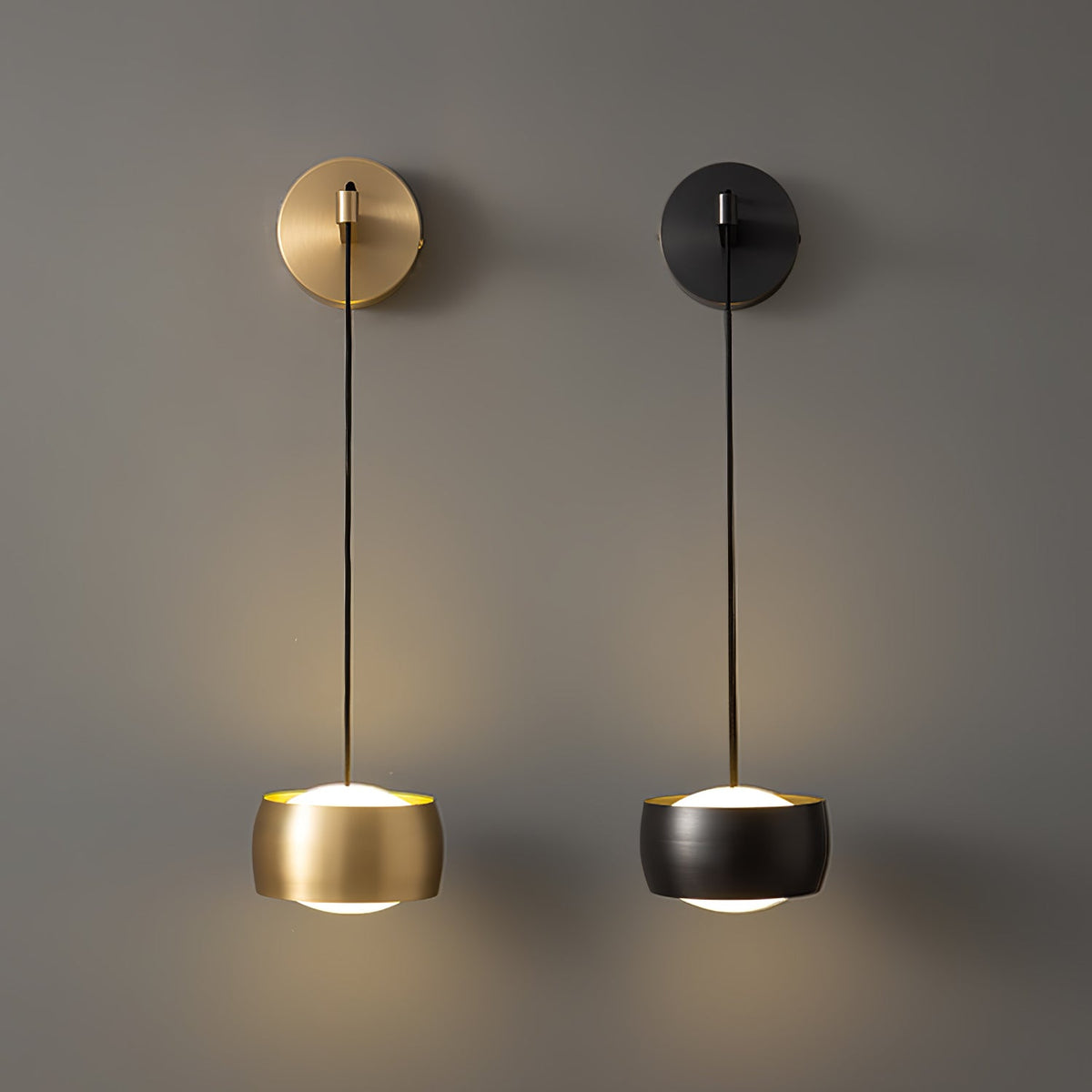 A brass Clérachic wall light and a black one side by side on a taupe wall, both lit from beneath their shades.