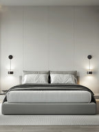 Pair of black Clérachic wall lights either side of a bed with grey bedding and a pale upholstered headboard.
