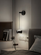 Lit black Clérachic wall light above a bed, with a shelf of books, a dried flower stem and wall sockets beside it.