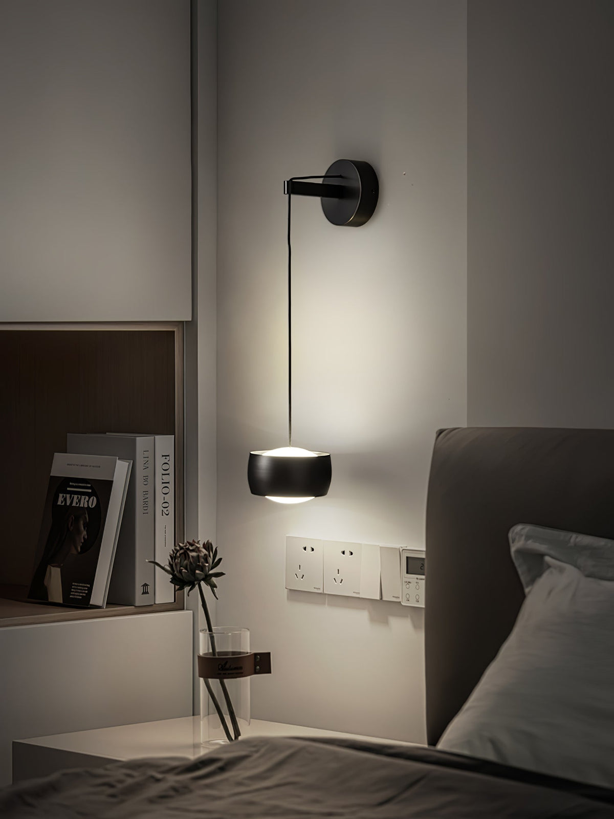 Lit black Clérachic wall light above a bed, with a shelf of books, a dried flower stem and wall sockets beside it.