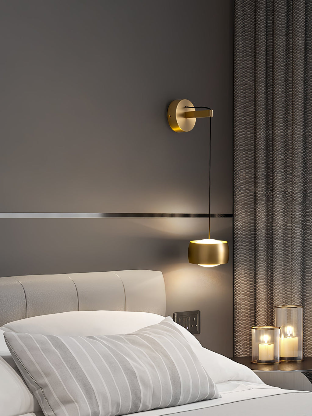 Brass Clérachic wall light lit above a bed with striped pillows, with a grey curtain and candles in glass holders at the right.