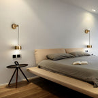 A pair of brass Clérachic wall lights either side of a low timber bed, with a round side table and an open book on the bed.