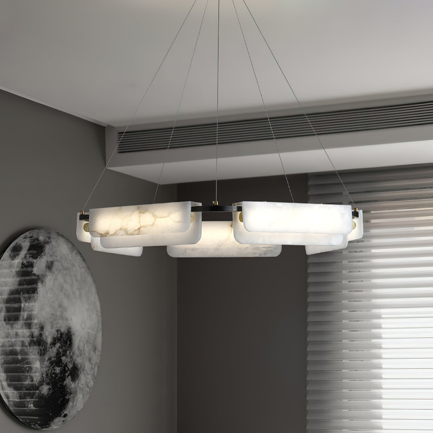 Clérachic Delia chandelier lit beside a large moon artwork, its alabaster panels glowing against a grey wall.