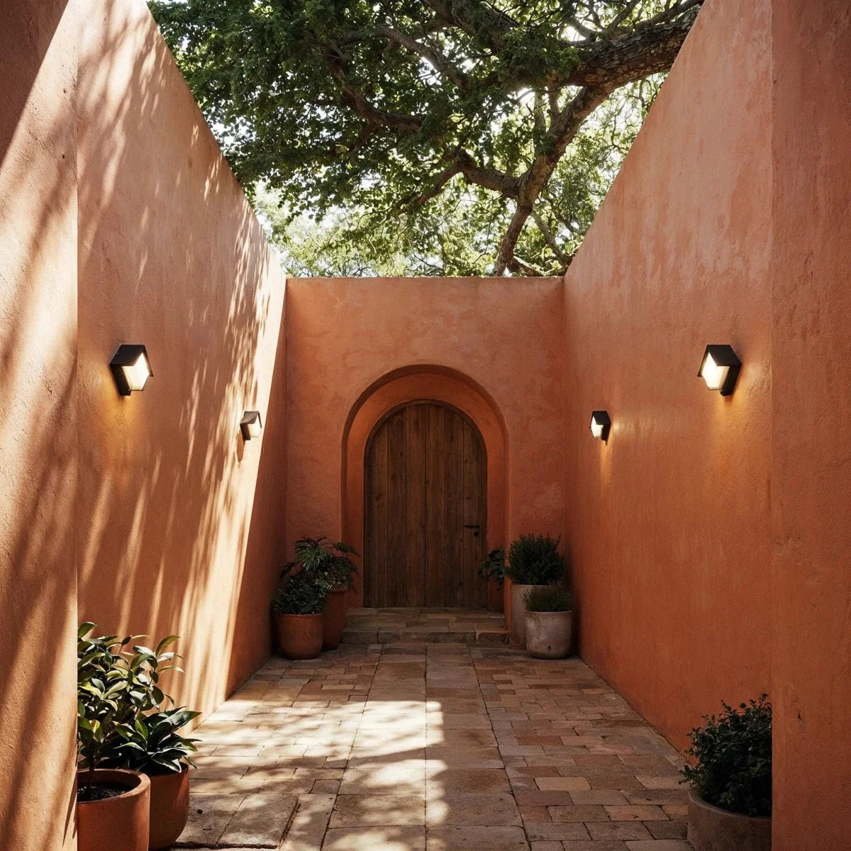 Clérachic compact black outdoor wall lamps glowing along terracotta courtyard walls on either side of an arched wooden door