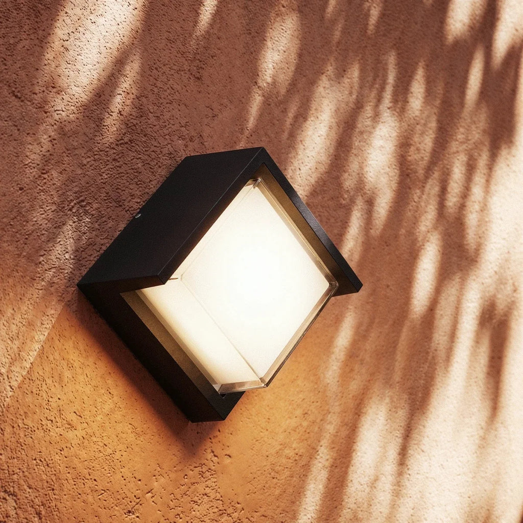 Close view of a Clérachic compact black outdoor wall lamp glowing warm white on a terracotta wall dappled with leaf shadows