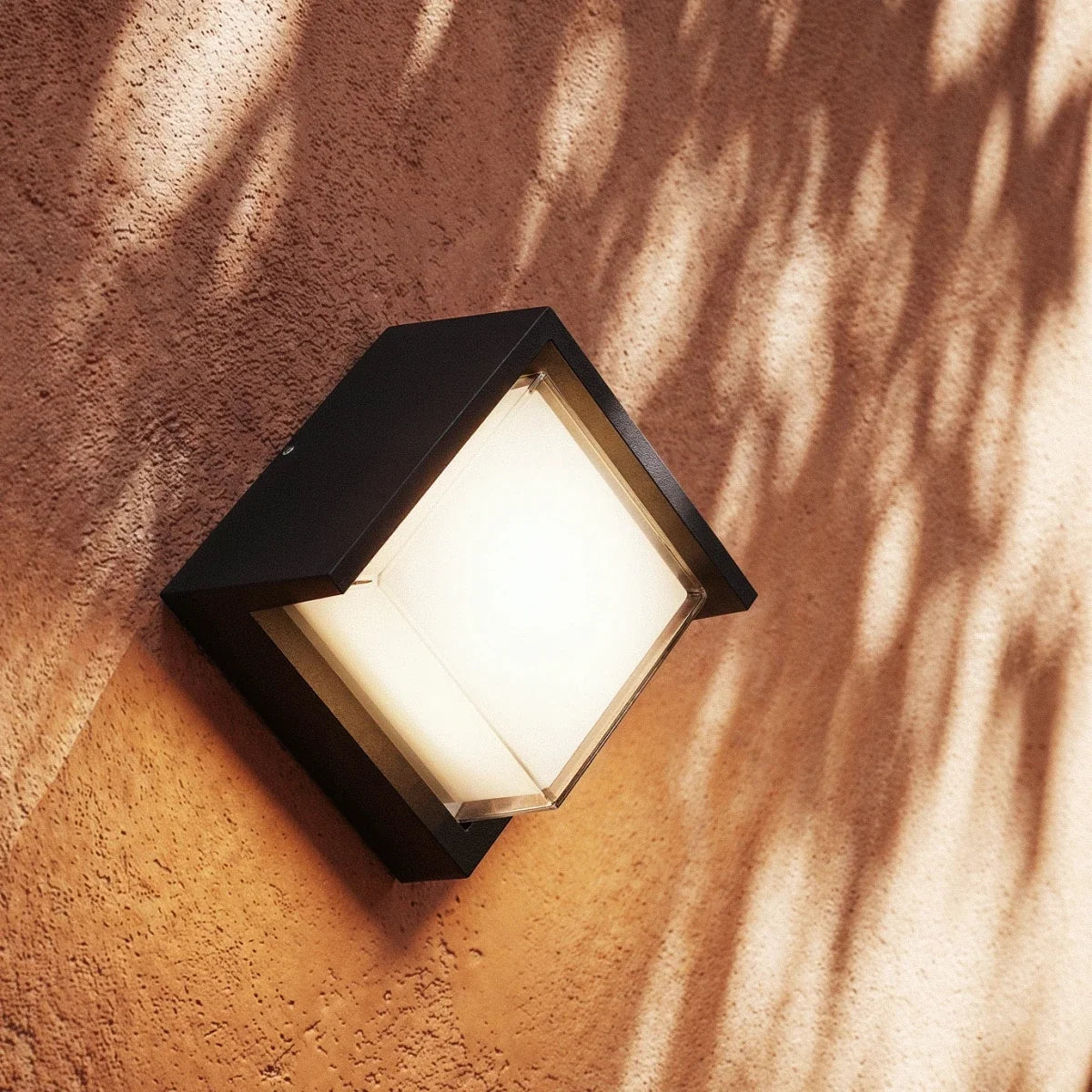 Close view of a Clérachic compact black outdoor wall lamp glowing warm white on a terracotta wall dappled with leaf shadows