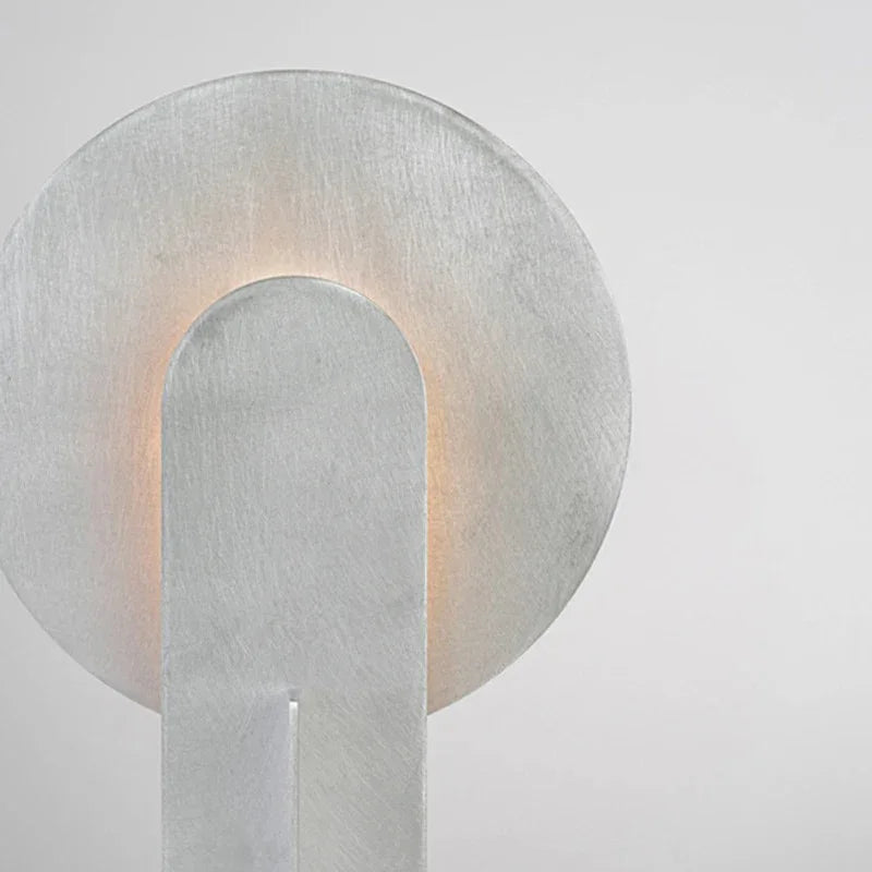 Clérachic disc-and-slab table lamp in close-up, showing the fine brush grain across the metal disc and the rounded top of the solid front slab.