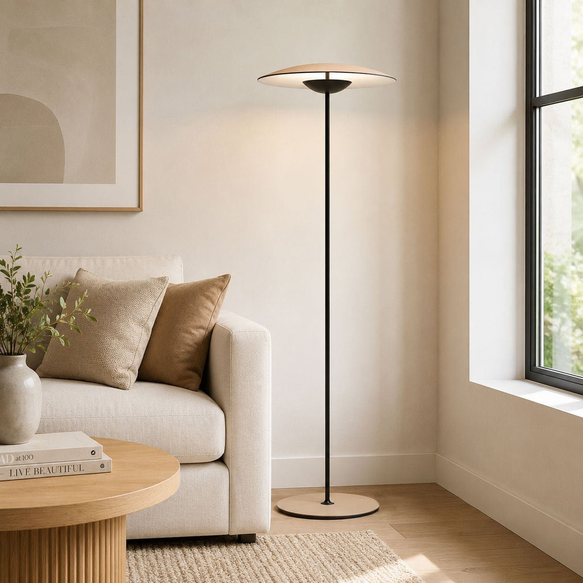A Clérachic floor lamp stands in a bright corner by a window, beside a cream sofa, cushions and a round fluted coffee table.