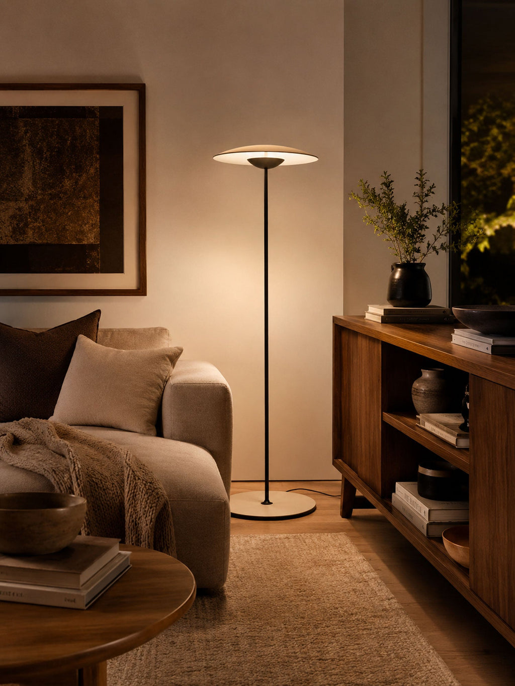 A Clérachic floor lamp glows in a dim, warm corner next to a lounge chair with a throw and a wooden shelving unit.