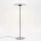 A Clérachic floor lamp with a wood-grain shade, black stem and round base, its cord trailing across a white floor.