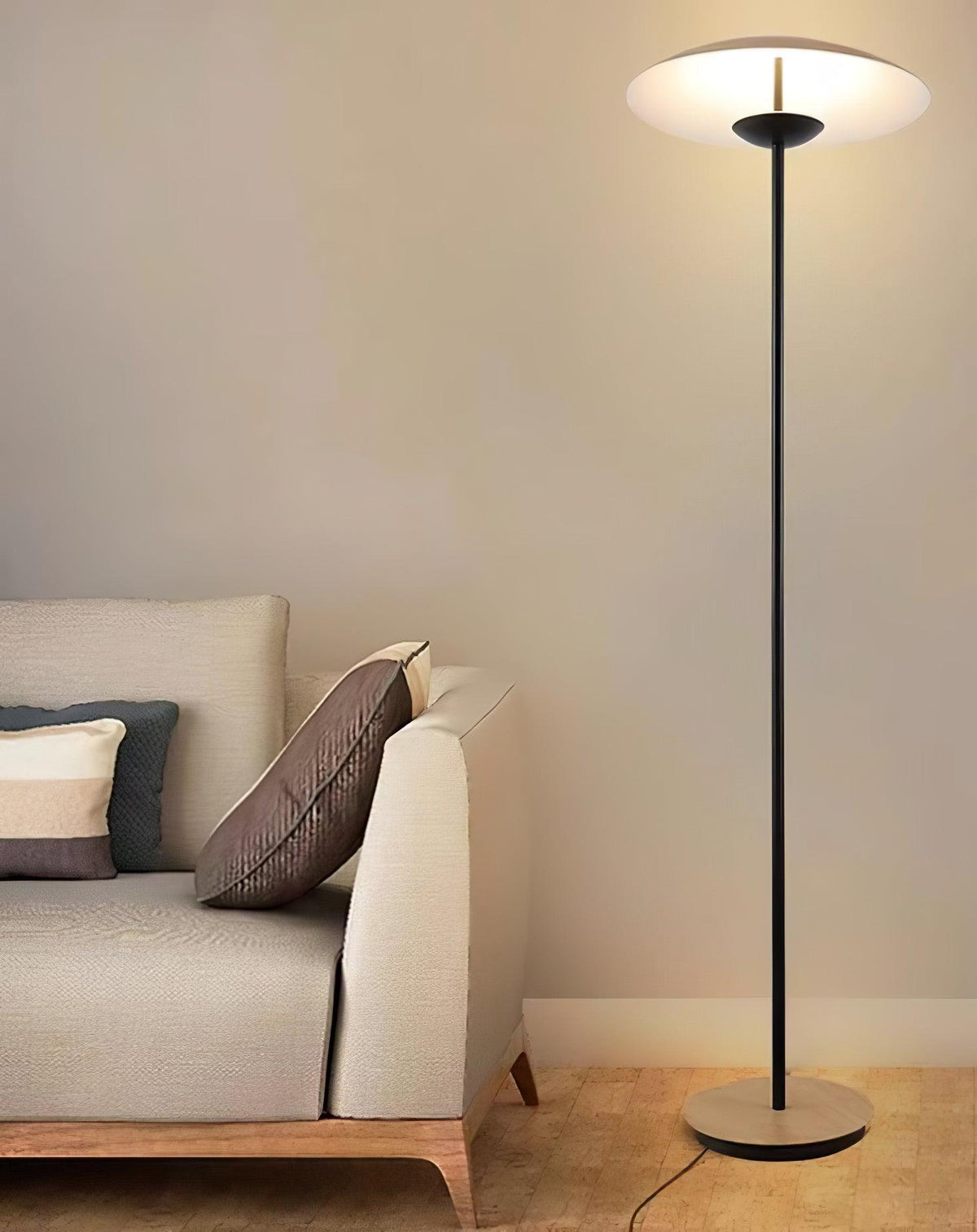 A Clérachic floor lamp lights a beige wall behind a cream sofa with striped cushions, its disc shade glowing white.